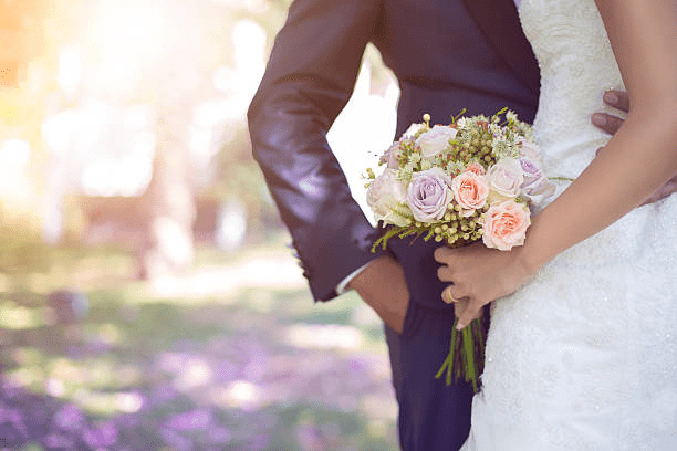 Bride in a white dress holding a bouquet of pink and white flowers stands next to a groom in a dark suit outdoors.