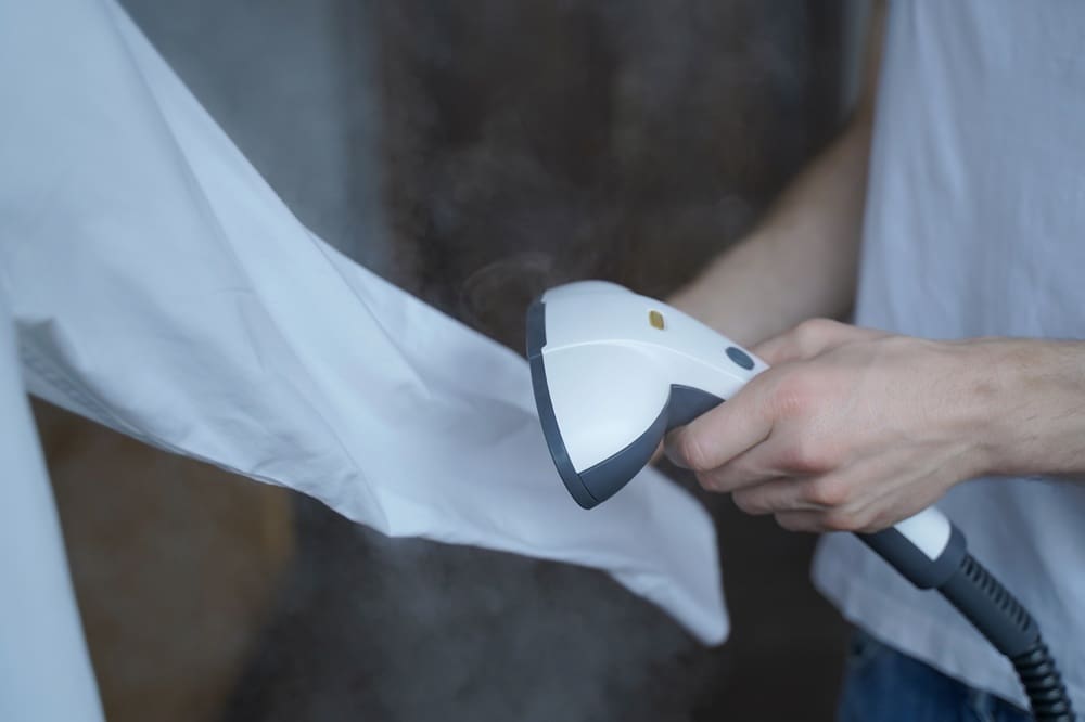 A person uses a handheld garment steamer to remove wrinkles from a white shirt.
