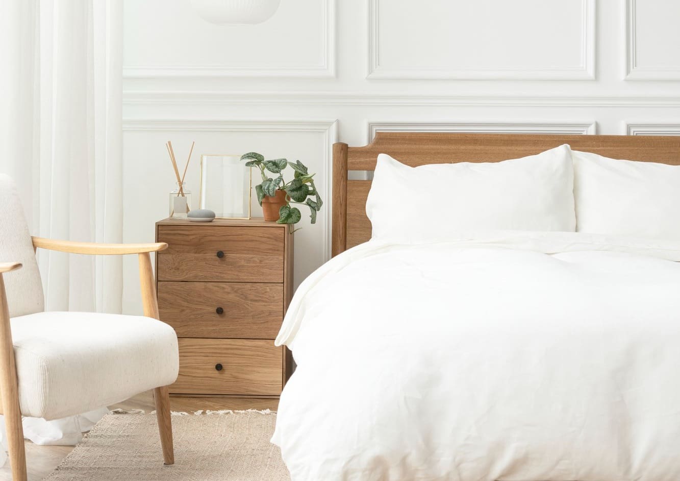A neatly made bed with white bedding is next to a wooden nightstand holding a plant, glass, and decor items. A beige armchair sits to the side in a bright, minimalist bedroom.