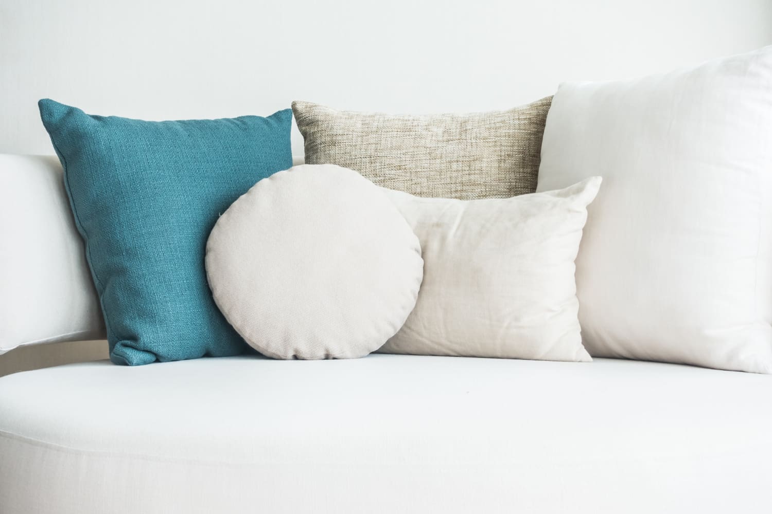 Four decorative pillows—one teal, two beige (one round), and one textured tan—arranged on a white curved sofa against a light background.