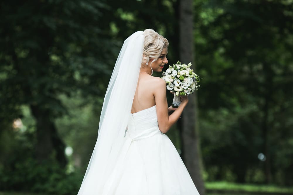 A bride in a white strapless gown and veil holds a bouquet of flowers while standing outdoors in a green, wooded area.