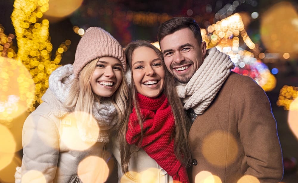 Three people wearing winter clothing smile and pose together outdoors at night with festive lights and blurred decorations in the background.