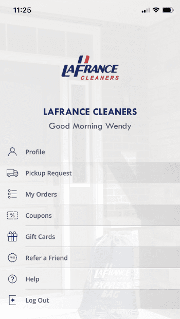 Mobile app home screen for LaFrance Cleaners showing user greeting, navigation menu options, and a blue laundry bag in the background.