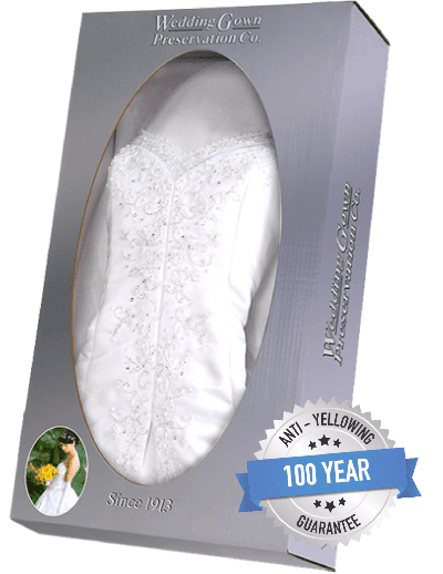 A white wedding gown with beaded details is displayed inside a grey preservation box labeled "Wedding Gown Preservation Co." with a "100 Year Anti-Yellowing Guarantee" badge.