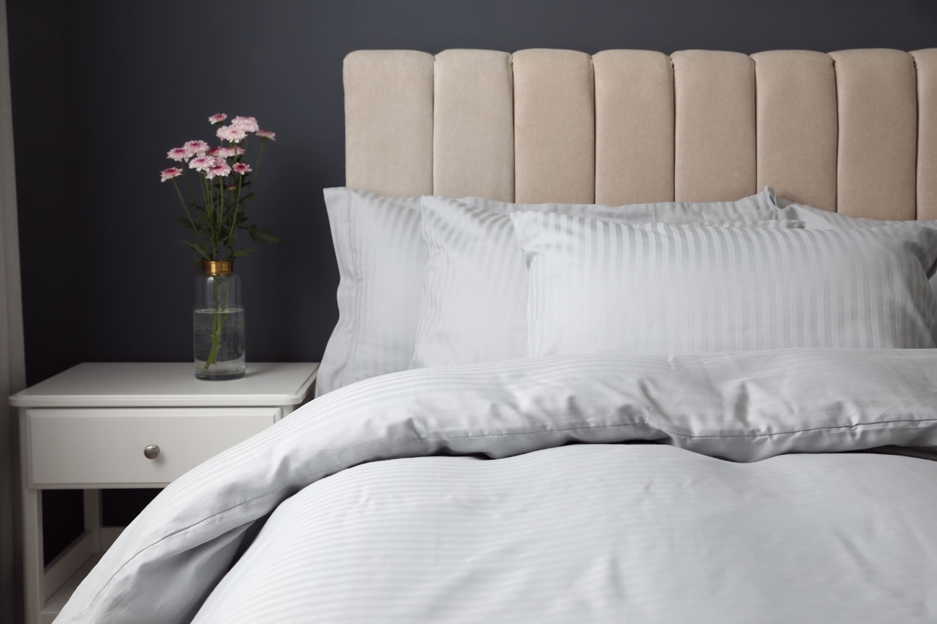 A neatly made bed with light gray striped bedding, a beige upholstered headboard, and a white nightstand holding a glass vase with pink flowers.