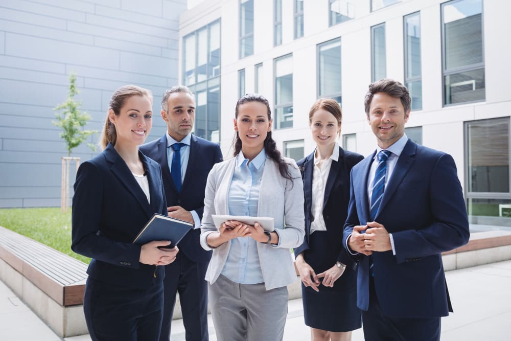 Five business professionals stand together outdoors in a modern office courtyard, dressed in formal attire and holding notebooks and a tablet.