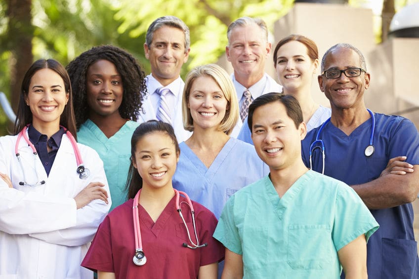 A diverse group of medical professionals, including doctors and nurses, stand together outdoors, smiling at the camera.