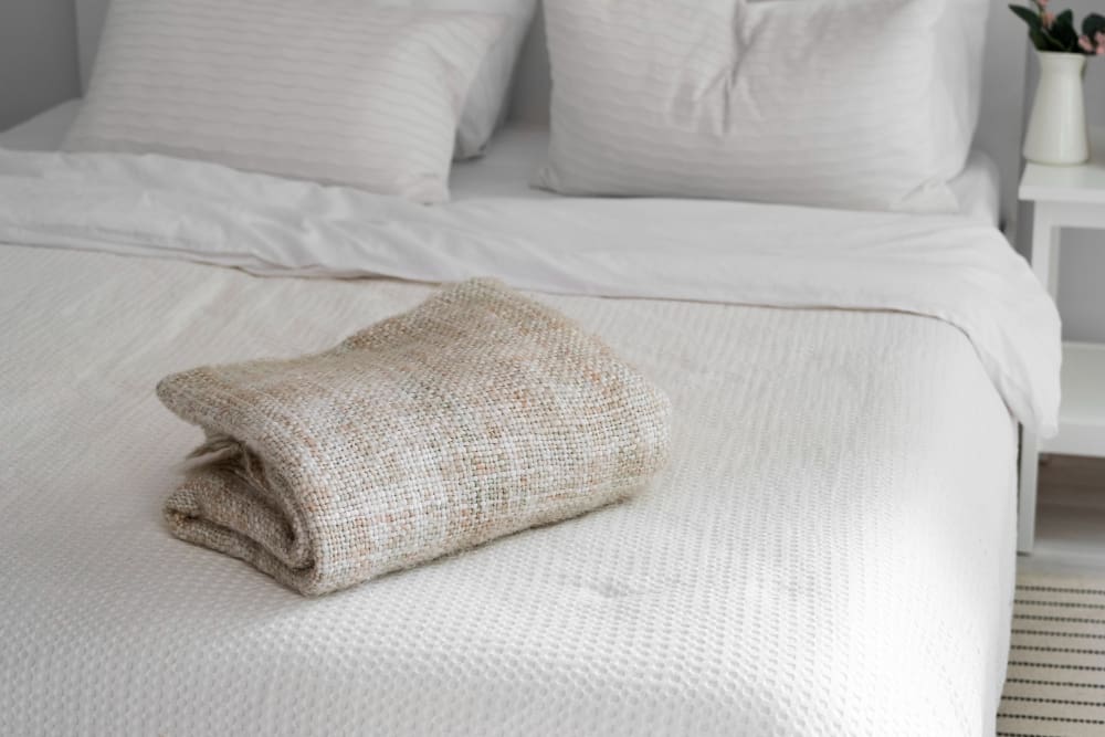 A neatly folded beige blanket sits on top of a made bed with white bedding and two pillows; a bedside table with a vase is visible in the background.