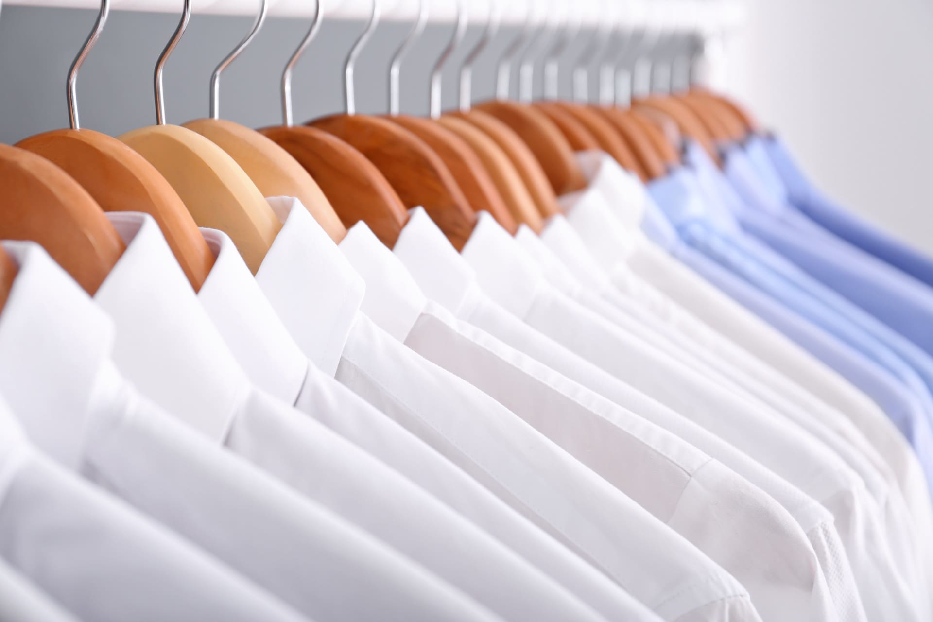 A row of neatly hung dress shirts on wooden hangers, mostly white with a few light blue shirts, displayed on a clothing rack.