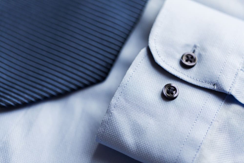 Close-up of a light blue dress shirt cuff with black buttons next to a dark, diagonally striped necktie.