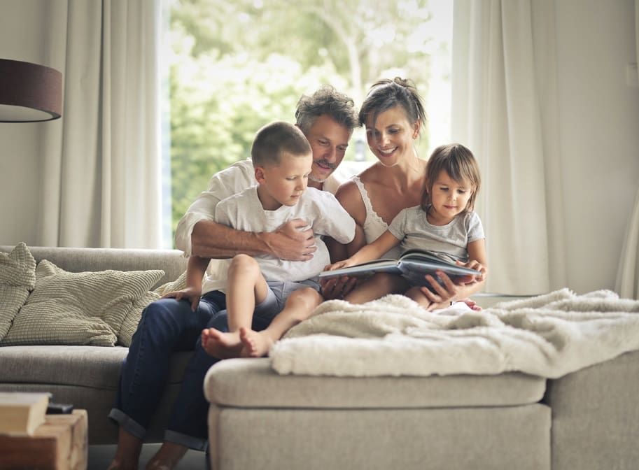 A family of four sits on a couch together, with the parents reading a book to their two young children in a bright living room.
