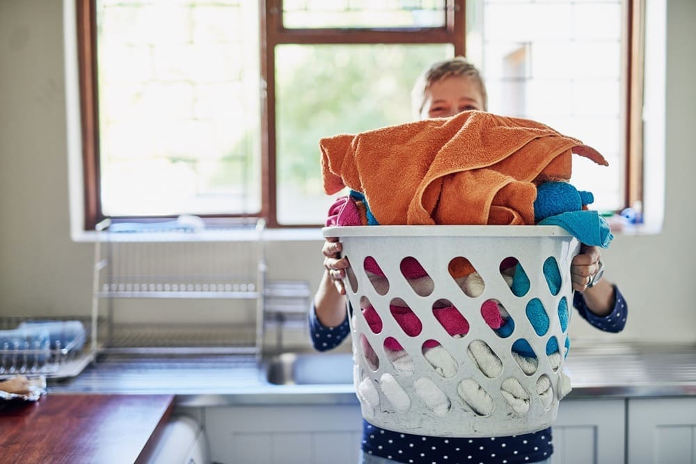 Person holding a laundry basket filled with colorful towels in a kitchen or laundry room, with a sink and drying rack in the background.