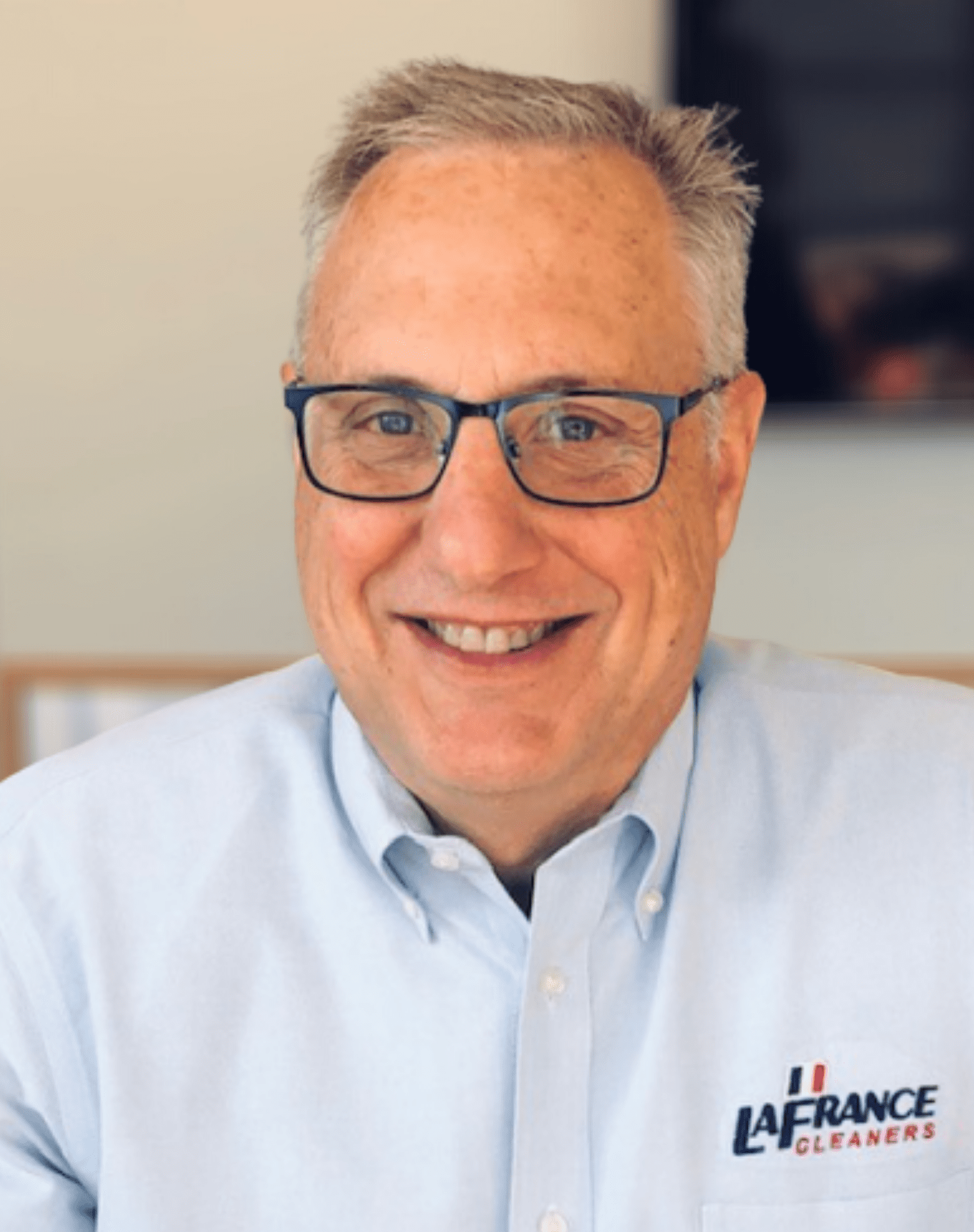 Smiling man wearing glasses and a light blue shirt with the "LaFrance Cleaners" logo, seated indoors against a blurred background.