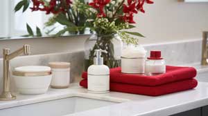 A bathroom countertop with a sink, soap dispenser, jars, two red towels, and a vase of red flowers in the background.