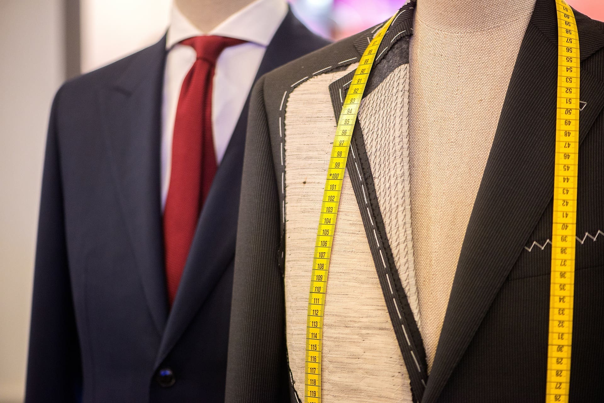 Two mannequins display suits; one finished with a red tie, the other partially tailored with exposed stitching and a yellow measuring tape draped around its neck.