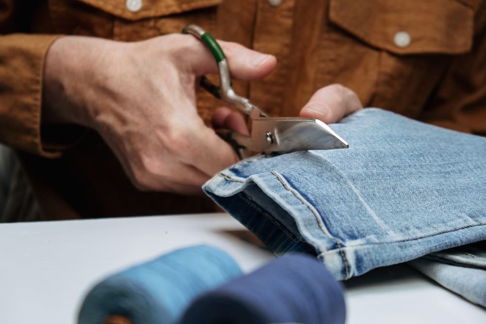 A person cuts a folded pair of blue jeans with scissors, while sewing threads are visible in the foreground.
