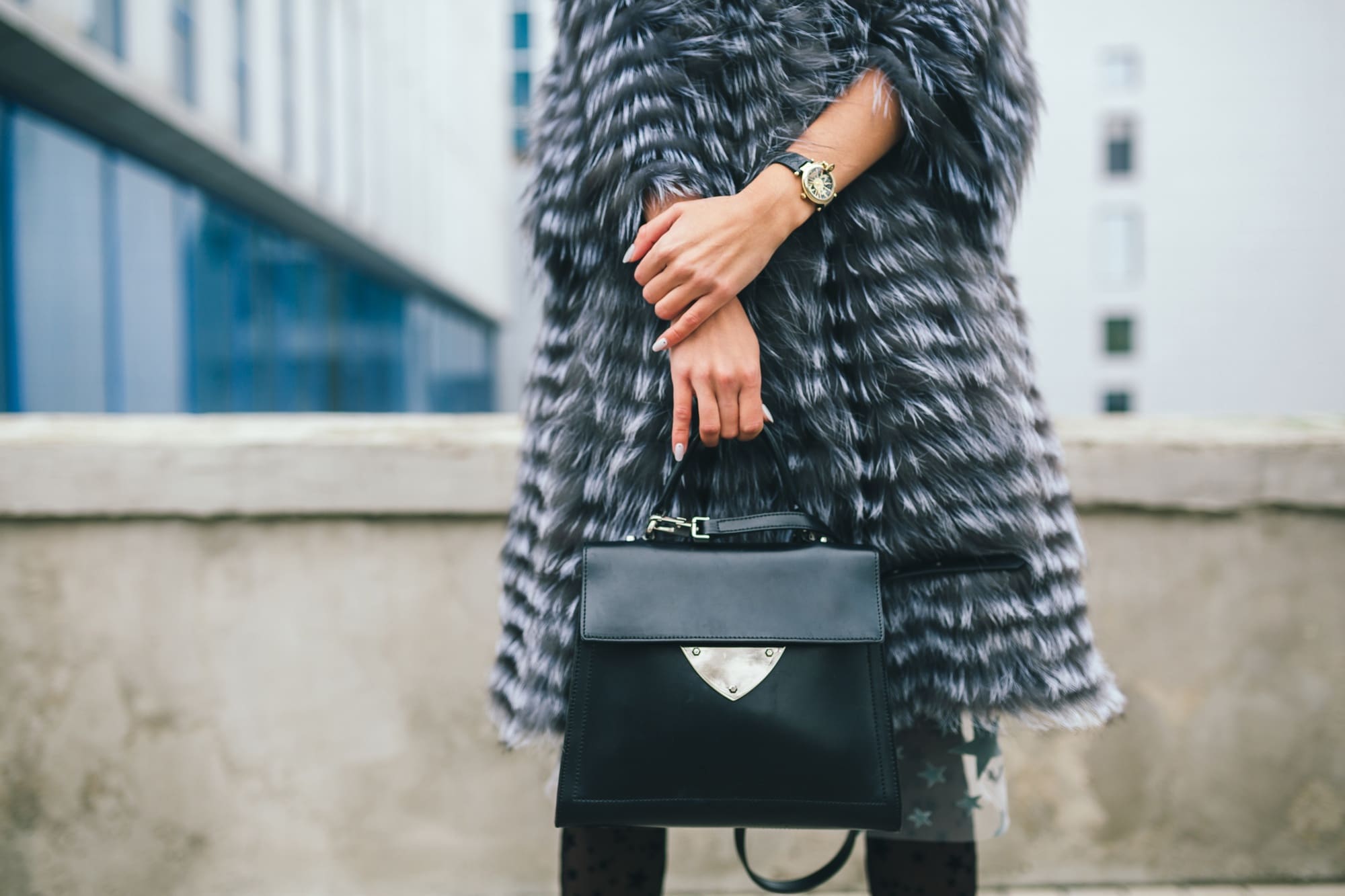 Person wearing a gray faux fur coat and wristwatch, holding a black handbag, standing outdoors near a concrete wall with buildings in the background.