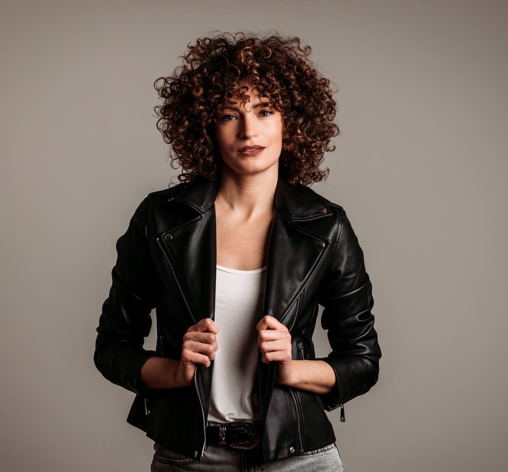 Woman with curly hair wearing a black leather jacket and white top, standing against a plain, neutral background.