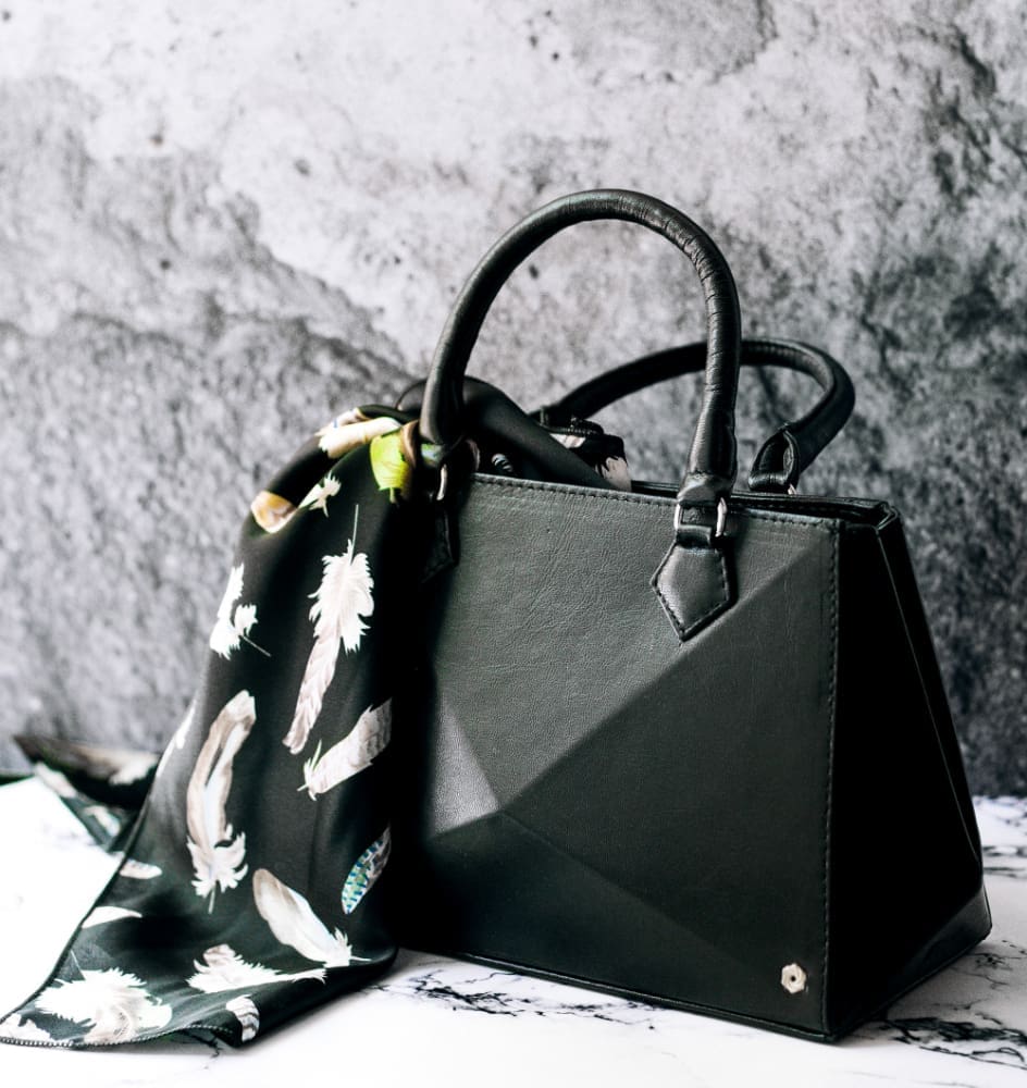 A black structured handbag with two handles is placed on a marble surface. A black scarf with white feather patterns is draped over one handle. Gray stone wall in the background.