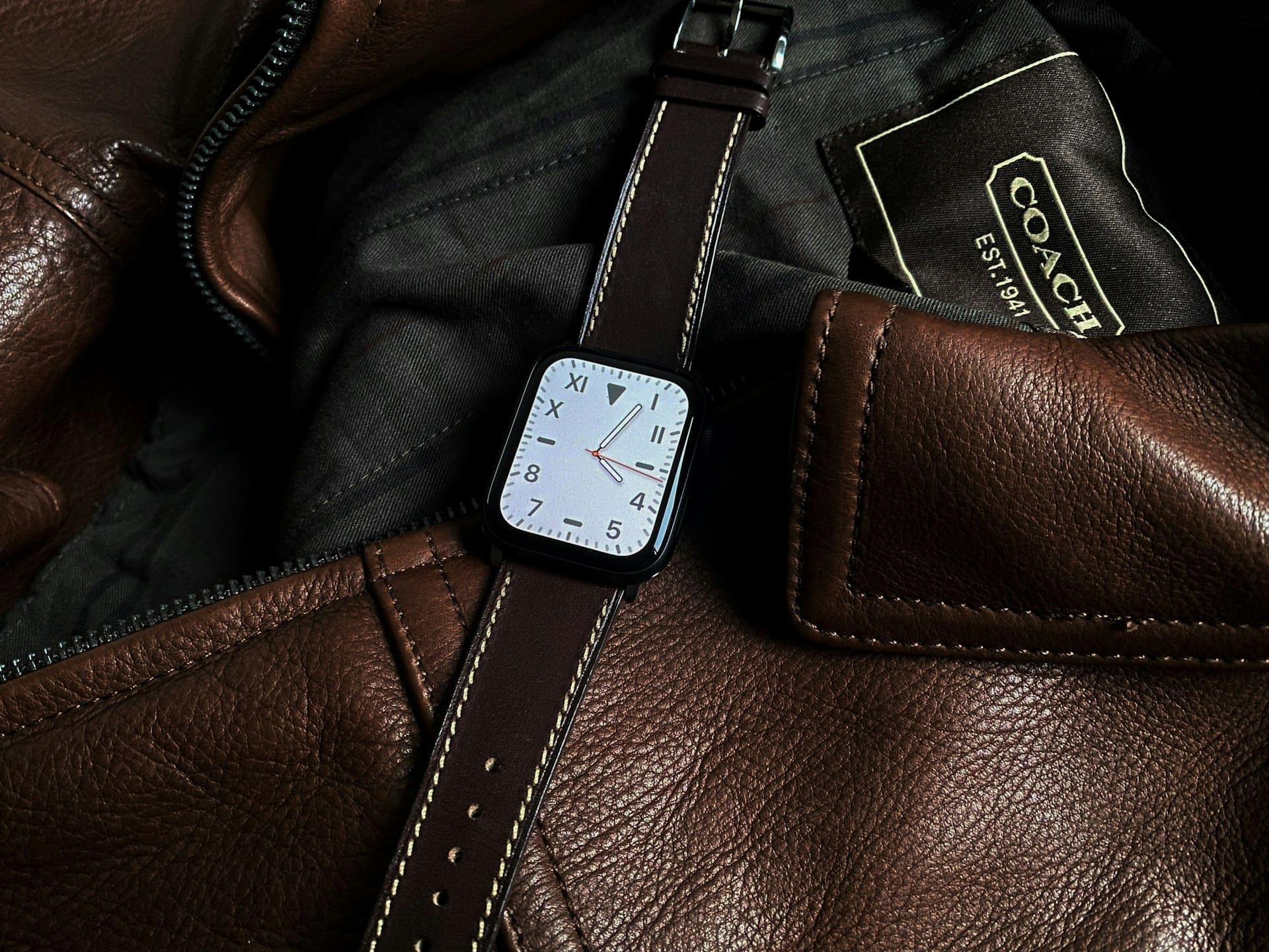 A smartwatch with a brown leather strap rests on a brown leather Coach jacket.
