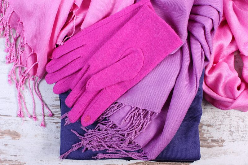 A pair of pink gloves rests on folded purple and pink scarves with fringe, placed on a light wooden surface.